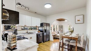 A modern kitchen with white cabinets, a marble countertop, and a dining area with a wooden table and chairs. - 🌿 SoCo Retreat by Creek with Workspace | Creekside Bungalow 🎶 (Austin)