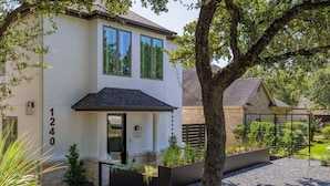 Et moderne hus i to etager med hvid facade, nummereret 1240, med en veranda, et stort træ og en velholdt have med forskellige planter samt en grusbelagt indkørsel.