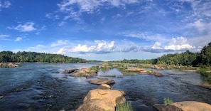 Un río con rocas y vegetación, un cielo despejado y árboles a lo lejos.