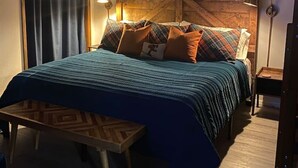 Un dormitorio con cama, cabecera de madera, mesita de noche, lámpara, un cuadro en la pared y una ventana con cortinas.