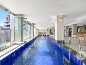 A modern indoor swimming pool with clear glass walls, a blue tiled floor, and a bench along the side. - 2-bed City Retreat With Gym and Pool Access (Adelaide)