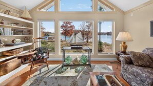 A spacious living room with a high ceiling, large windows offering a view of a lake and sailboat, a wooden rocking chair, a patterned sofa, and a bookshelf filled with books and decorative items. - Waterfront Lake Anna Private Side Retreat + Boathouse Oasis (Mineral)