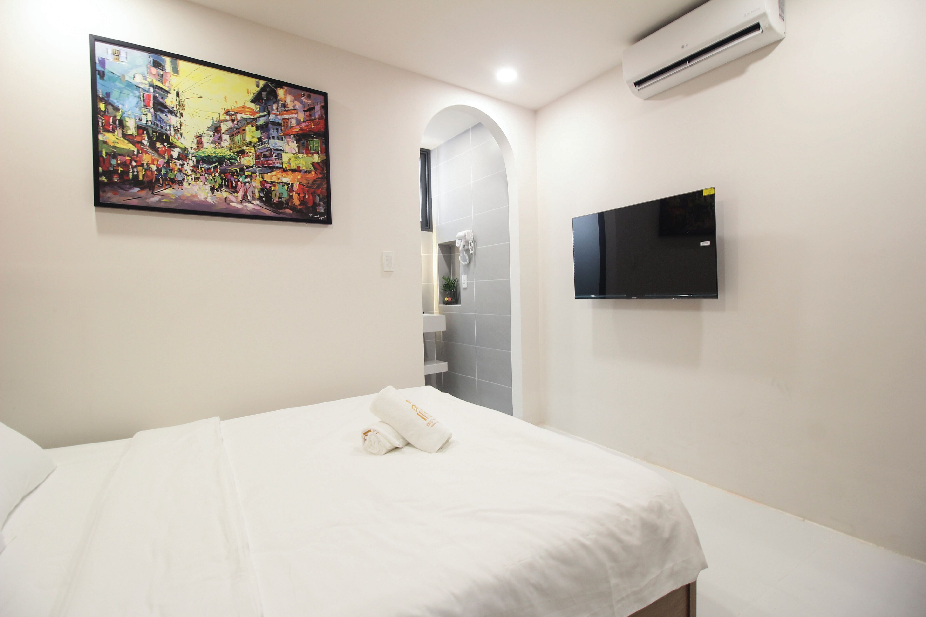 A hotel room with a bed, a painting, a TV, and an air conditioning unit.