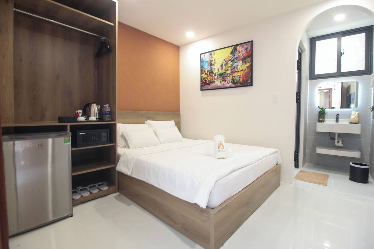 A modern hotel room with a wooden bed, a built-in wardrobe, a small kitchenette, and a bathroom with a mirror and sink.