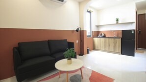 A modern living room with a black sofa, a small round table with a plant, and a kitchen area in the background. - De Lustora Hotel & Apartment - District 1, HCM. (Ho Chi Minh City)