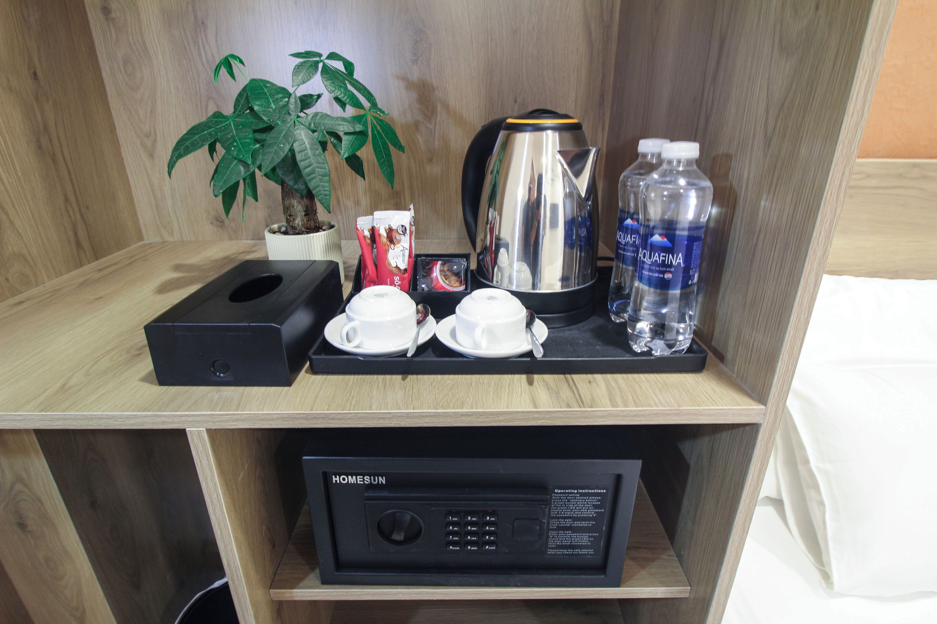 A hotel room with a wooden desk, a coffee maker, two cups, a kettle, a water bottle, a plant, and a control panel.