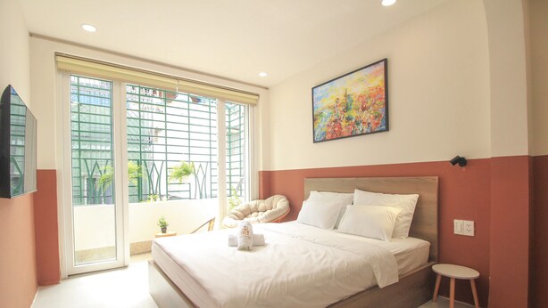 A bedroom with a large bed, a window with a view of buildings, a painting on the wall, and a small table. - De Lustora Hotel & Apartment - District 1, HCM. (Ho Chi Minh City)