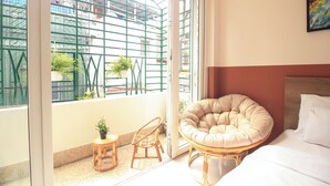A room with a bed, a round cushioned chair, a small table with a plant, and a view of buildings through a window with a metal grid. - De Lustora Hotel & Apartment - District 1, HCM. (Ho Chi Minh City)