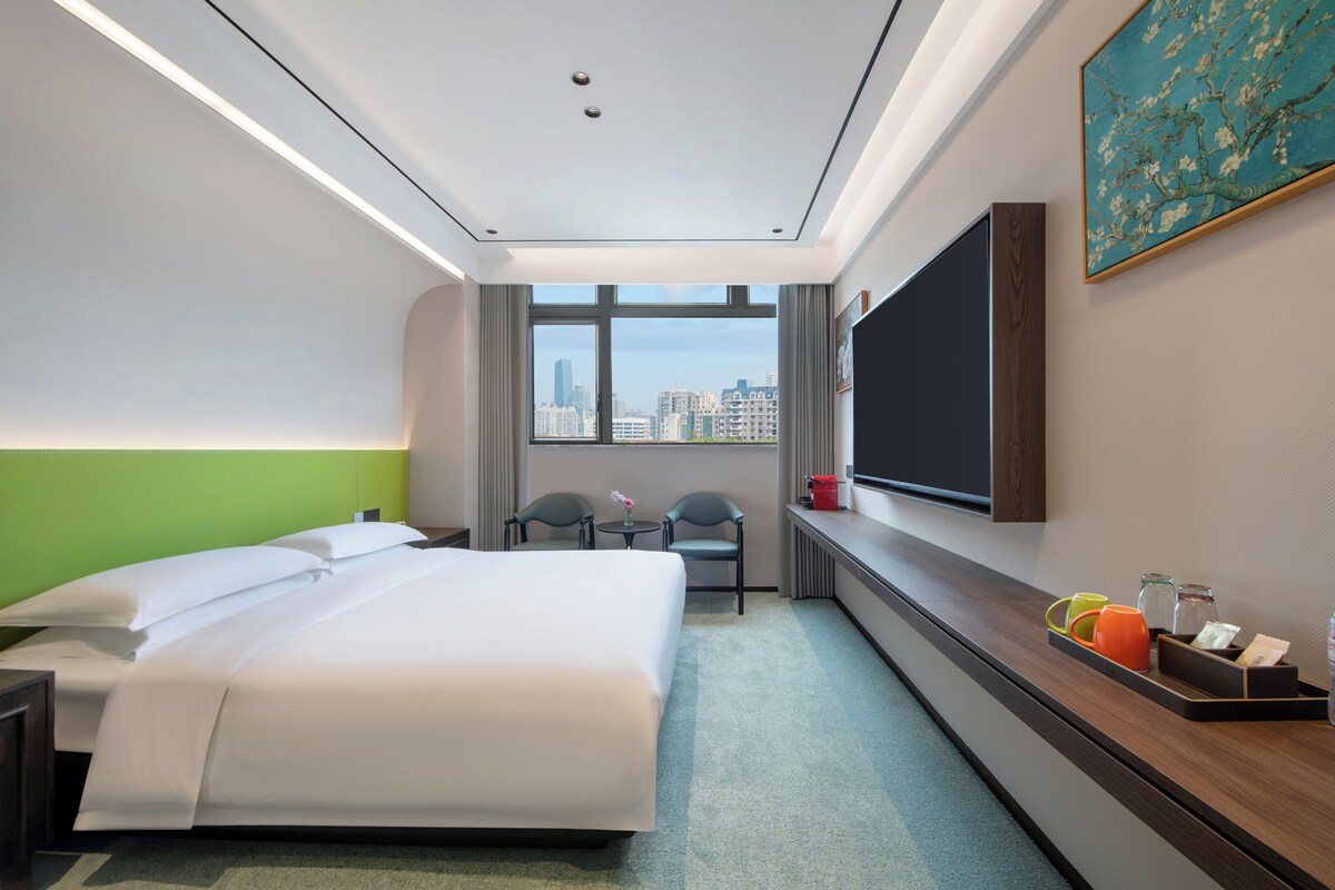 A hotel room with a large bed, a desk, a chair, a TV, and a window with a city view.