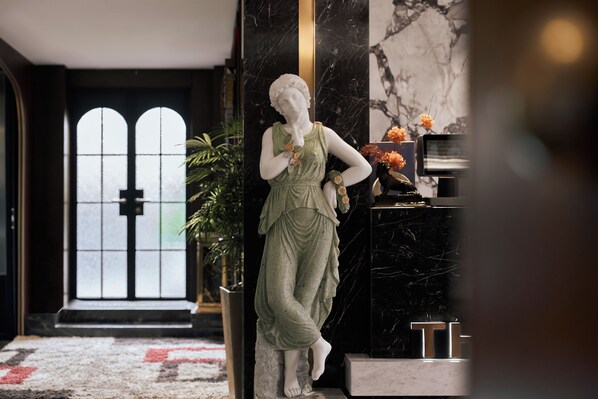 A statue of a woman in a green dress standing in a hallway with a marble floor, a black wall, and a door with a cross symbol.