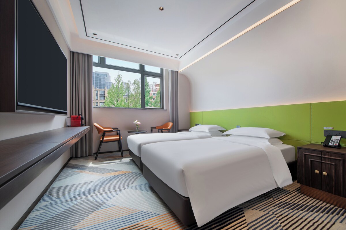 A modern hotel room with two beds, a desk, a chair, a small table with a vase, and a view of buildings through large windows.