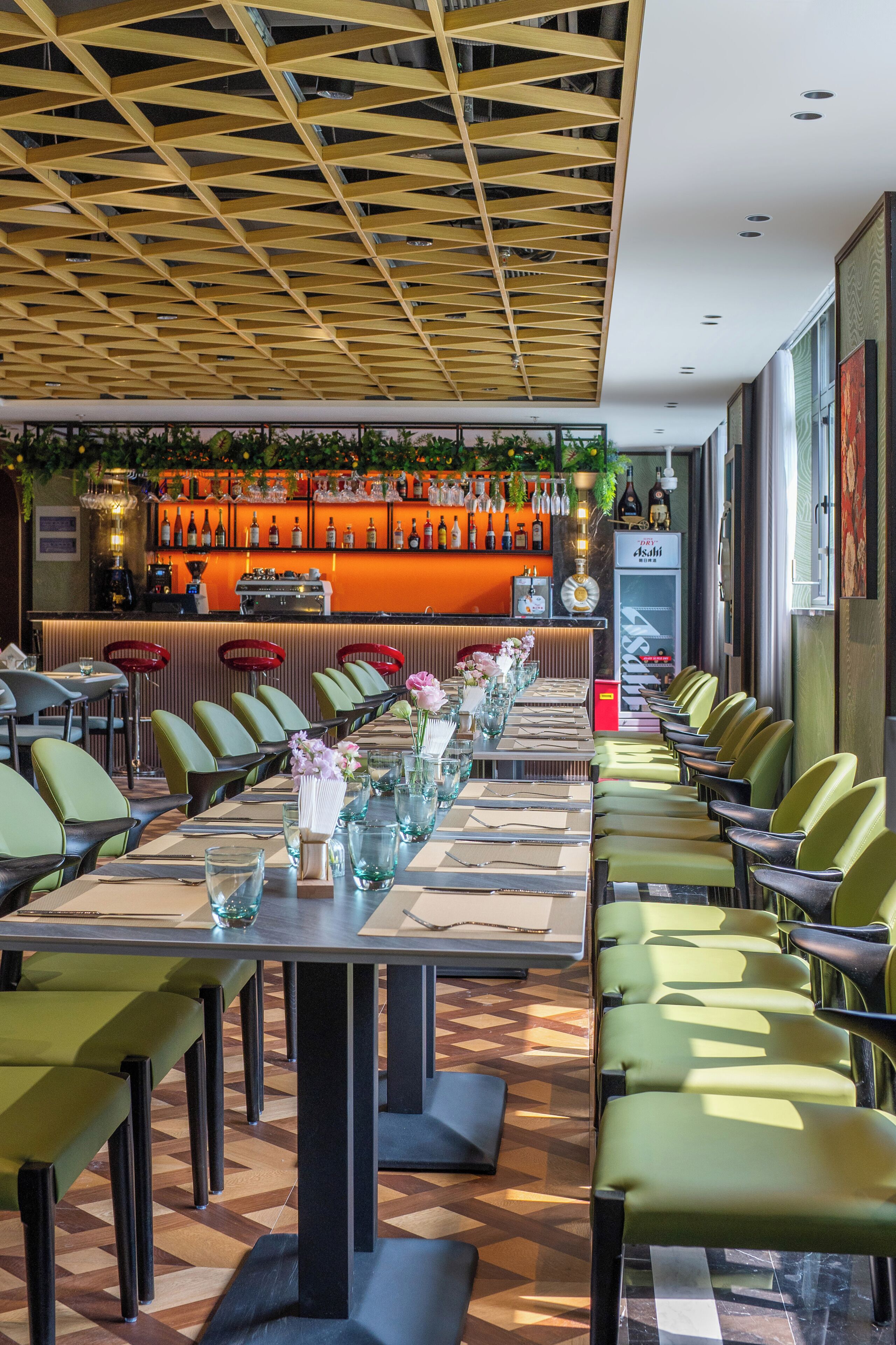 A restaurant with a long table set for dining, a bar area with bottles, and a modern ceiling design.