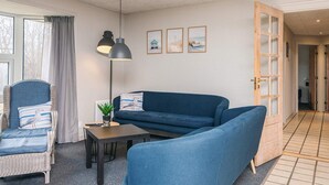 Un salón acogedor con un sofá azul, una butaca de mimbre, una mesita y una lámpara de pie. En la pared hay tres cuadros enmarcados y una puerta de madera que da a otra habitación.