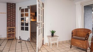 Una habitación con suelo de baldosas, una silla de mimbre, una mesita con una planta y una puerta entreabierta que da a otra estancia.