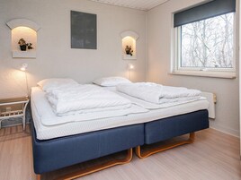 Cama doble con estructura azul oscuro y ropa de cama blanca, dos lámparas de pared, una ventana con vistas a árboles y una mesita de noche con lámpara.