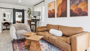 Un soggiorno moderno con un divano in pelle marrone, una poltrona beige, un tavolino in legno e un tappeto decorato. Alle pareti si trovano dipinti astratti e una scrivania con una lampada.