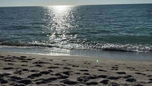 Een strand met voetafdrukken die naar het water leiden, een vliegende vogel erboven en de zon die in de zee weerkaatst.