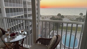 Een balkon met een glazen tafel, twee stoelen en uitzicht op zee bij zonsondergang.