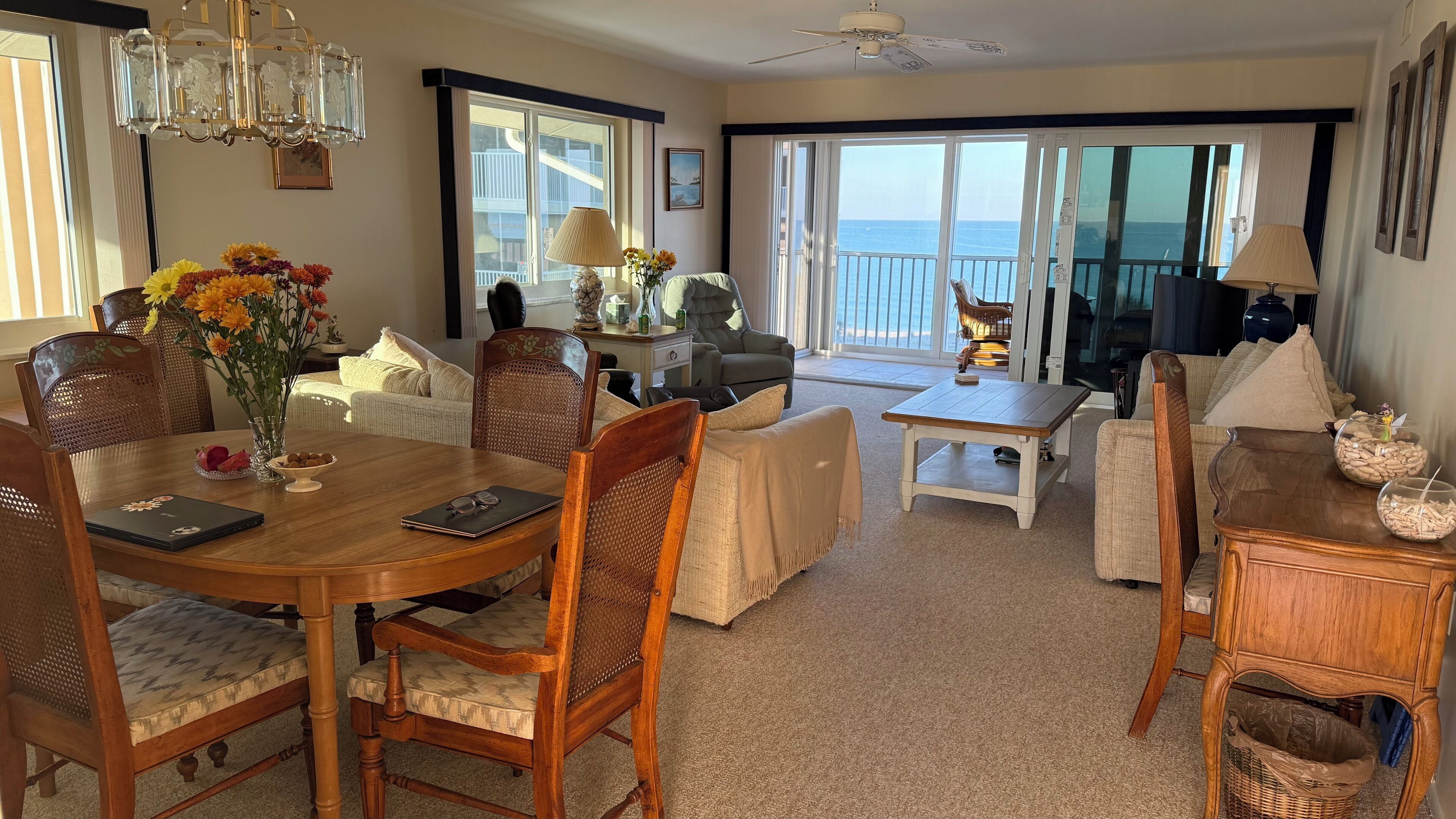 A spacious living room with a dining area, large windows with a view of the sea, a chandelier, and a ceiling fan.