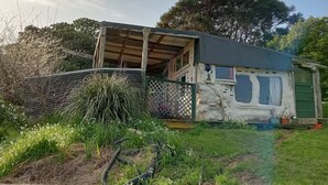 A rustic house with a wooden porch, a green fence, and a garden area with various plants. - The Mud House (Raglan)