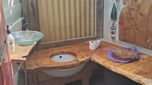 A rustic bathroom with a wooden sink, a green plastic container, and a window with a view of trees. - The Mud House (Raglan)