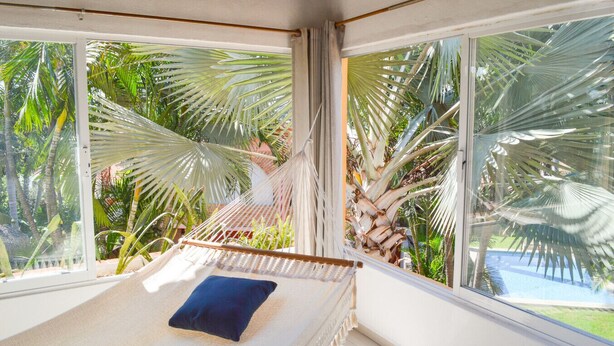 Eine Hängematte mit dunklem Kissen, große Fenster und Blick auf Palmen und einen Pool.