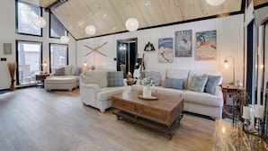 A spacious living room with a high ceiling, wooden beams, and large windows. There is a sofa set, a coffee table, and decorative items on the walls. - Double home escape in the trees (Angel Fire)