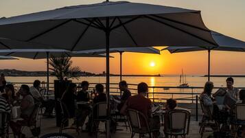 Zona de comedor al aire libre con mesas y sillas bajo sombrillas, con vista a un cuerpo de agua con barcos y un atardecer de fondo.