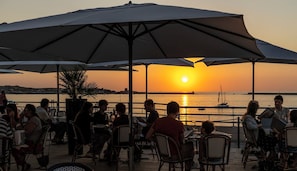 An outdoor dining area with tables and chairs under umbrellas, overlooking a body of water with boats and a sunset in the background. - Thalazur St Jean de Luz (Saint-Jean-de-Luz)