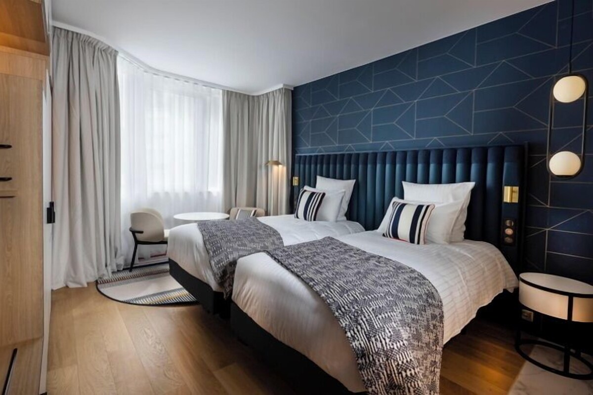 A hotel room with two beds, a round table, a chair, and a large window with curtains.