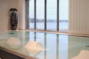 Ein Hallenbad mit großen Glaswänden, einer Vase mit Blumen und Blick auf ein Gewässer durch Panoramafenster.