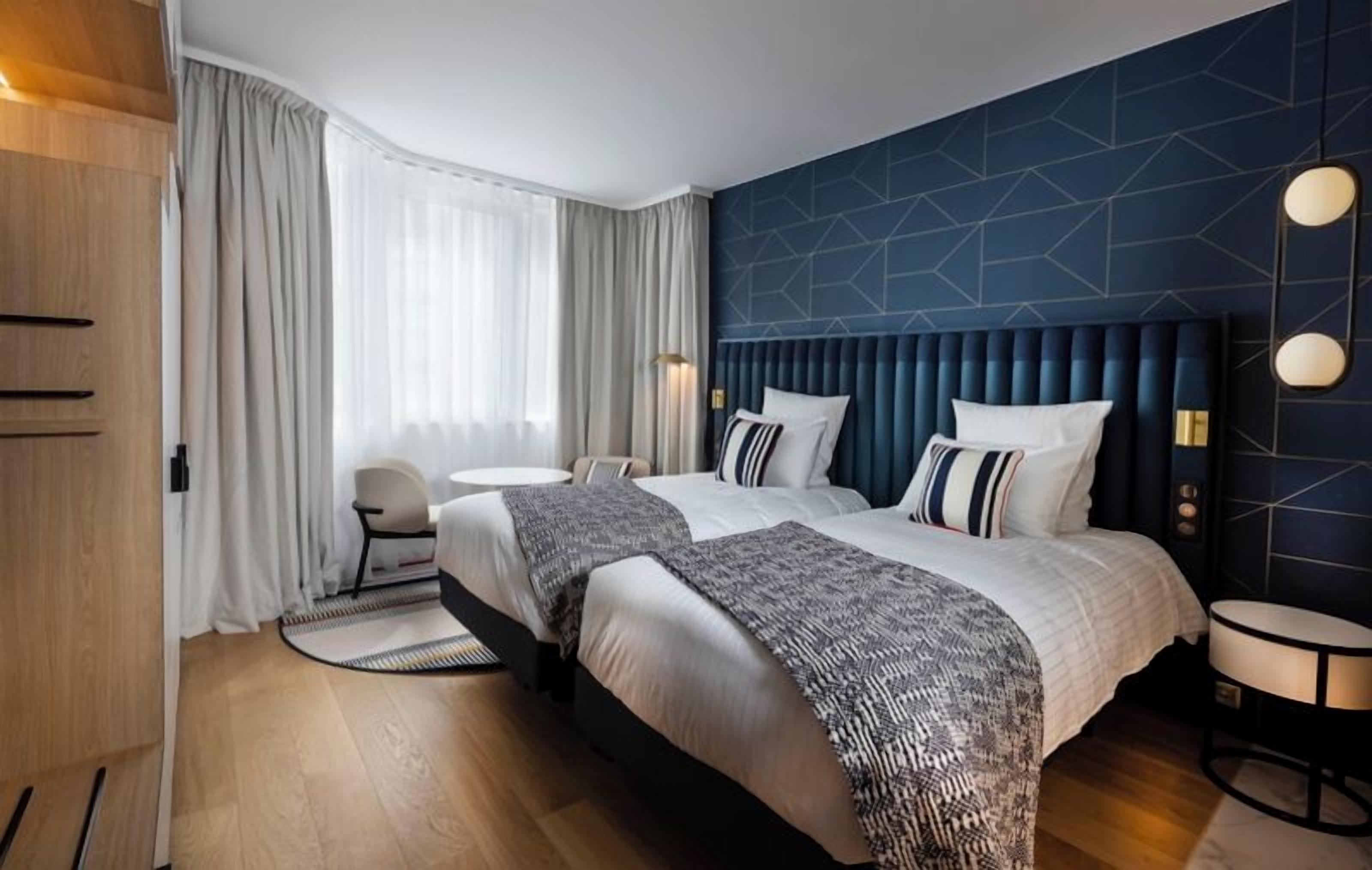 Habitación de hotel con dos camas, una mesa redonda, una silla y un gran ventanal con cortinas.