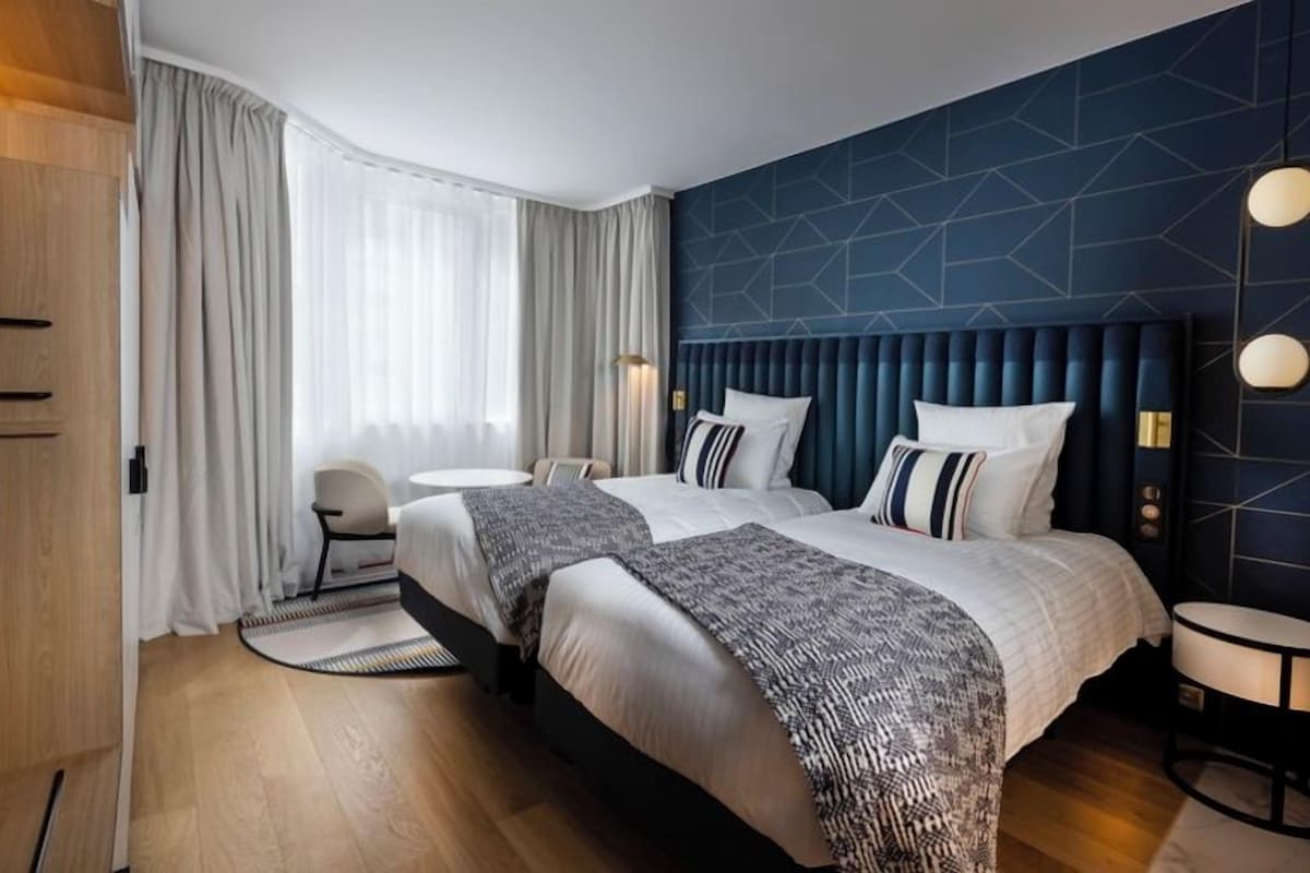 A hotel room with two beds, a round table, a chair, and a large window with curtains.
