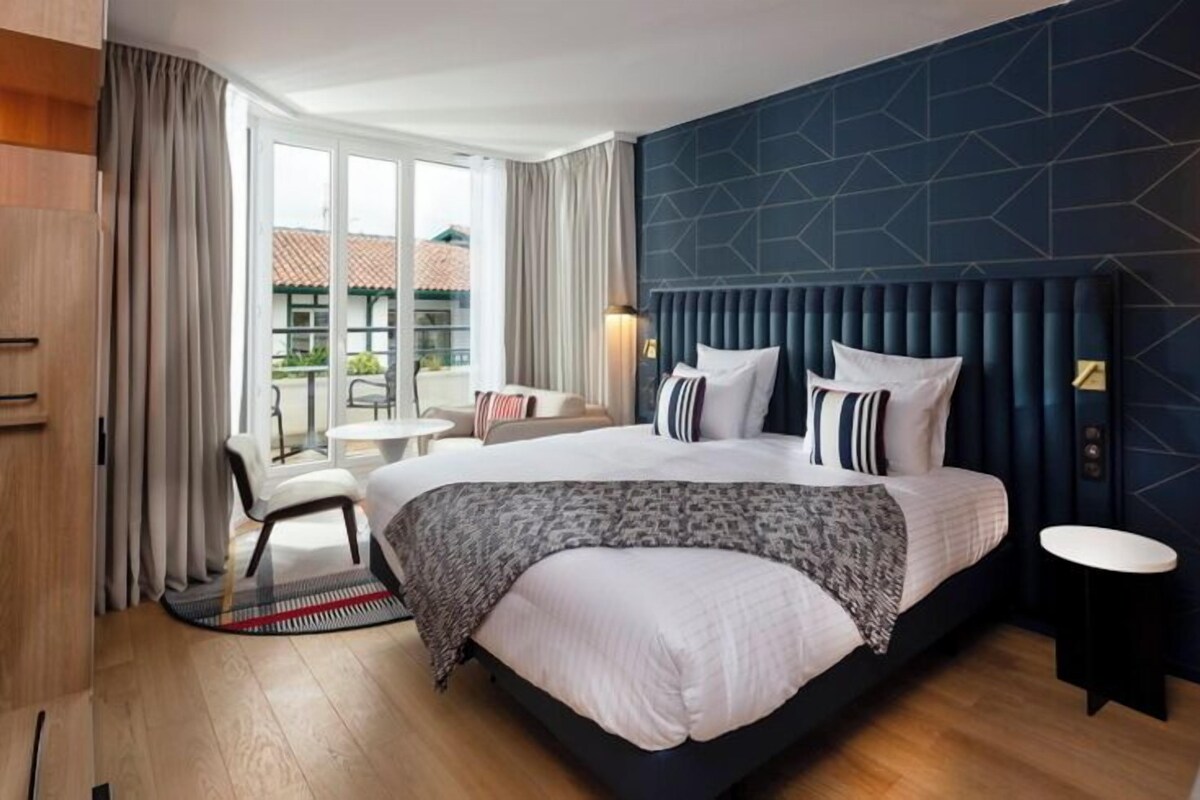 A modern hotel room with a large bed, a seating area with a chair and table, and a view of the outdoors through the window.