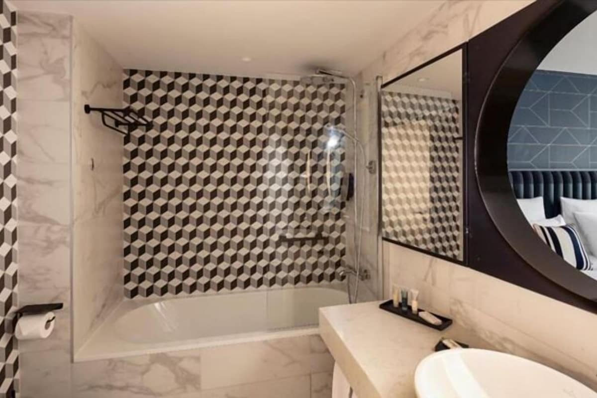 A bathroom with a geometric patterned shower wall, a round mirror, and a marble-tiled floor.