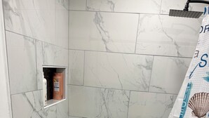 Een badkamer met marmeren tegels, een douchegordijn met een strandthema en een klein plankje met toiletartikelen.