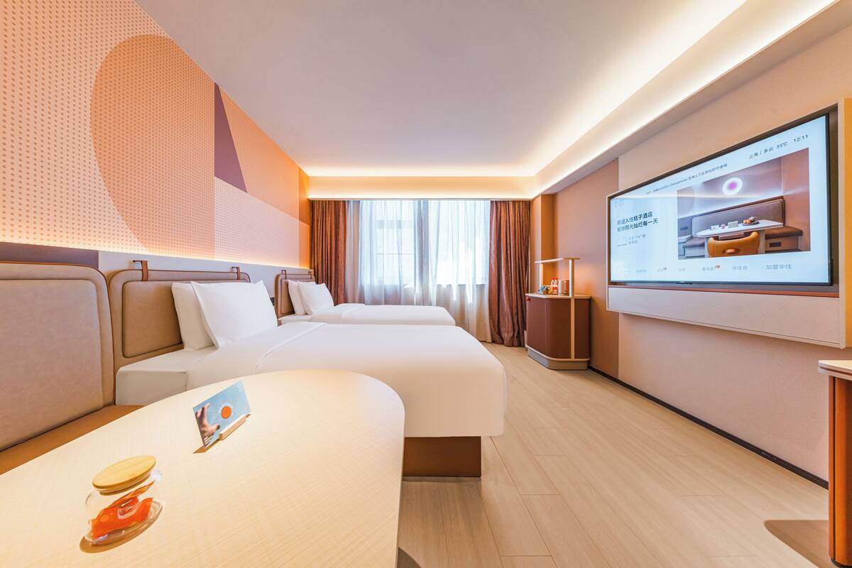 A hotel room with two beds, a large screen displaying a room layout, and a wooden floor.