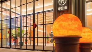 Entrada de un hotel con amplias ventanas, plantas en macetas y un letrero con caracteres chinos.