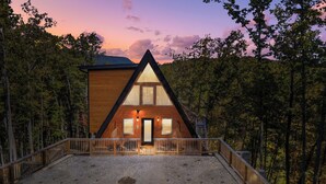 Una cabana de fusta moderna, de forma triangular, amb un porxo frontal i una entrada de grava, envoltada per un bosc dens.