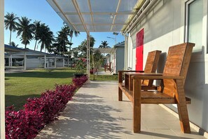 A covered outdoor seating area with wooden benches, a pathway lined with red plants, and a view of palm trees and buildings in the background. - Saga Village Hotel & Resort Bintan By Zuzu (Bintan)
