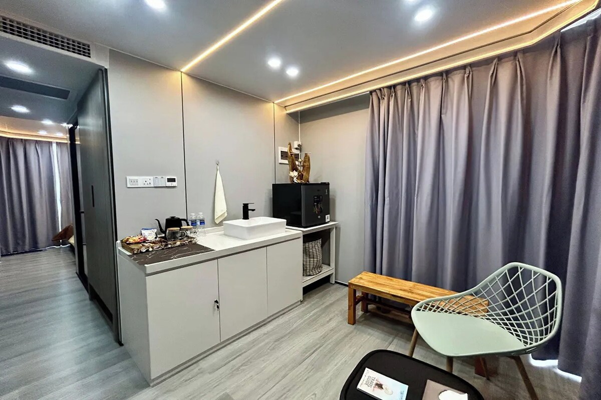 A modern hotel room with a kitchenette, a wooden bench, a white chair, and a large window with curtains.