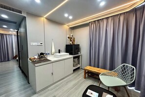 A modern hotel room with a kitchenette, a wooden bench, a white chair, and a large window with curtains. - Saga Village Hotel & Resort Bintan By Zuzu (Bintan)