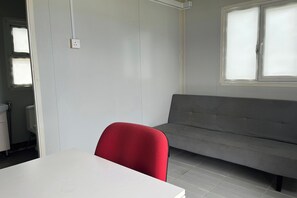 A room with a white table, a red chair, a grey sofa, and a window with blinds. - Saga Village Hotel & Resort Bintan By Zuzu (Bintan)