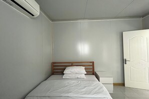 A small, neatly made bed with a wooden headboard in a room with light-colored walls and a closed door. - Saga Village Hotel & Resort Bintan By Zuzu (Bintan)
