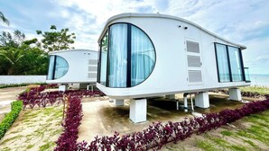 Deux structures modernes, blanches, dotées de toits incurvés et de grandes fenêtres, sont implantées dans un espace paysager agrémenté de feuillage violet.