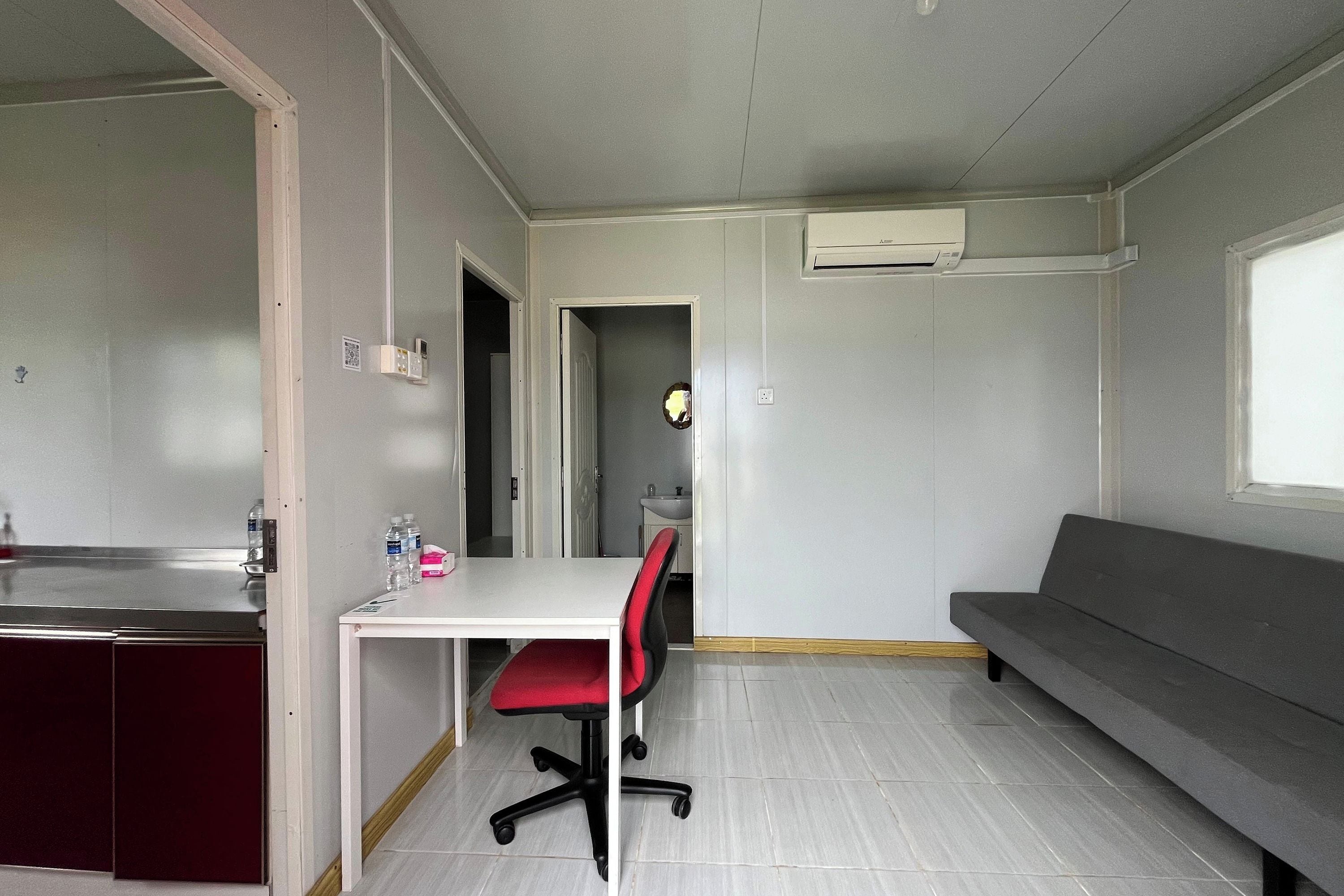 A compact room with a kitchenette, a dining table with a red chair, a grey sofa, and a small bathroom visible through an open door.