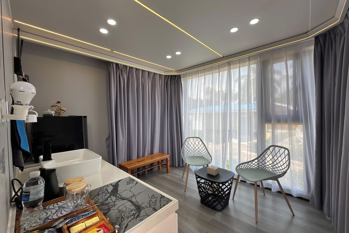 A modern room with a wooden bench, two chairs, a small table, a coffee maker, and a TV on a stand.