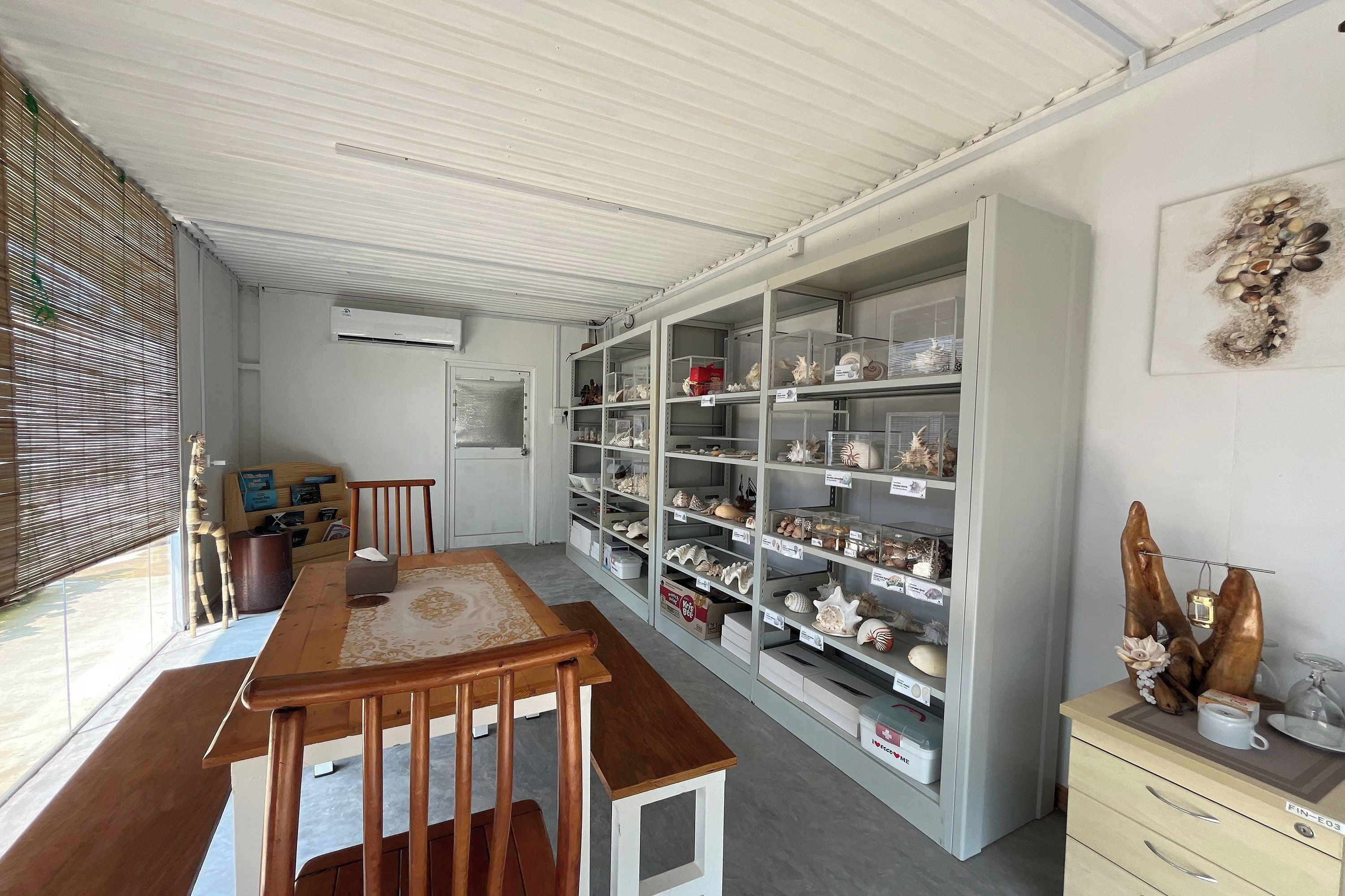 A room with a wooden dining table, chairs, a white cabinet with shelves, and a window with blinds.