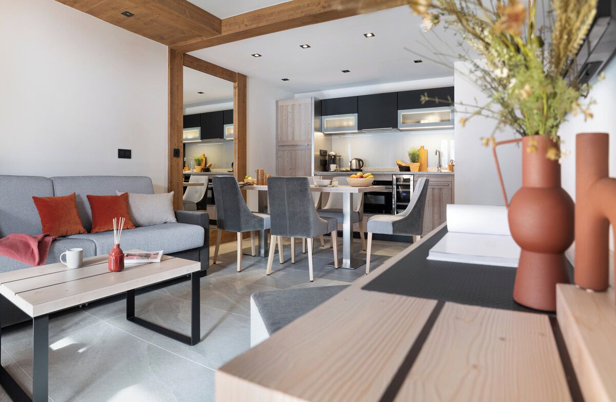 A modern living room with a sofa, coffee table, and chairs. A kitchen is visible in the background with cabinets and appliances.