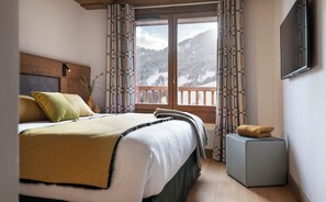 A bedroom with a bed, a bedside table, a TV, and a view of mountains through a window. - MGM Hôtels et Résidences - Hotel Laska (Les Contamines-Montjoie)